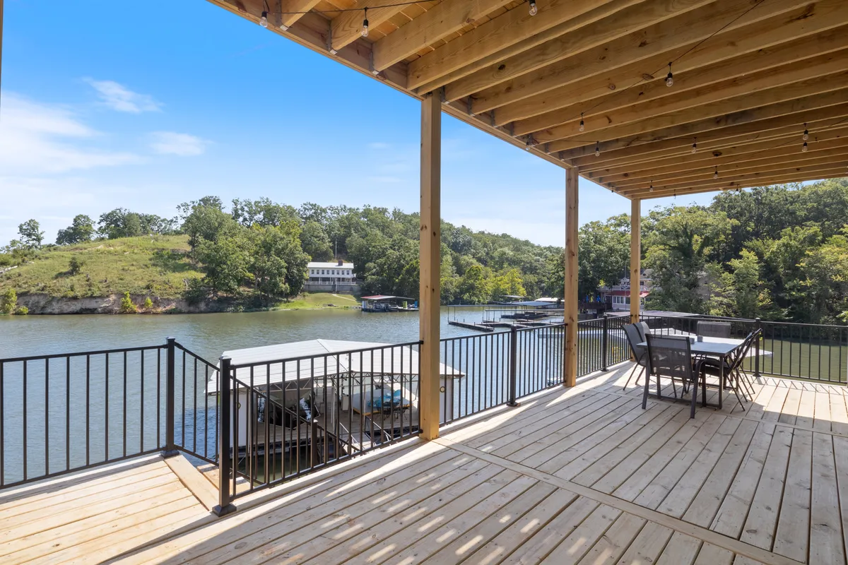A spacious deck with a sturdy railing offers wide views of the lake and is surrounded by trees. The area is set up for outdoor dining and lounging, providing a serene setting for morning coffee, afternoon relaxation, or sunset celebrations.
