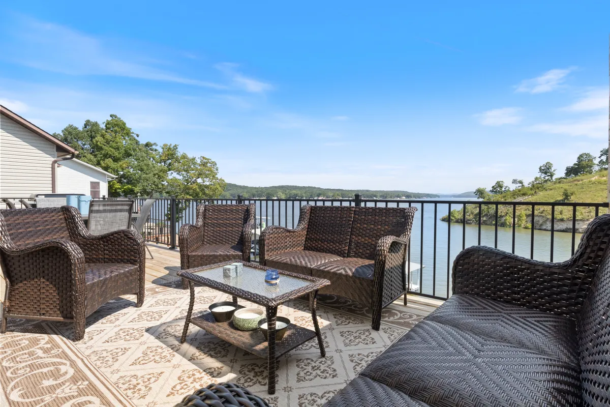A cozy outdoor lounge area with wicker furniture and a coffee table, set atop a plush decorative rug on the deck. The seating is arranged to foster intimate conversation, all set against a stunning lake backdrop. The atmosphere is serene, with soft lighting and the lake's reflective surface creating a peaceful mood.