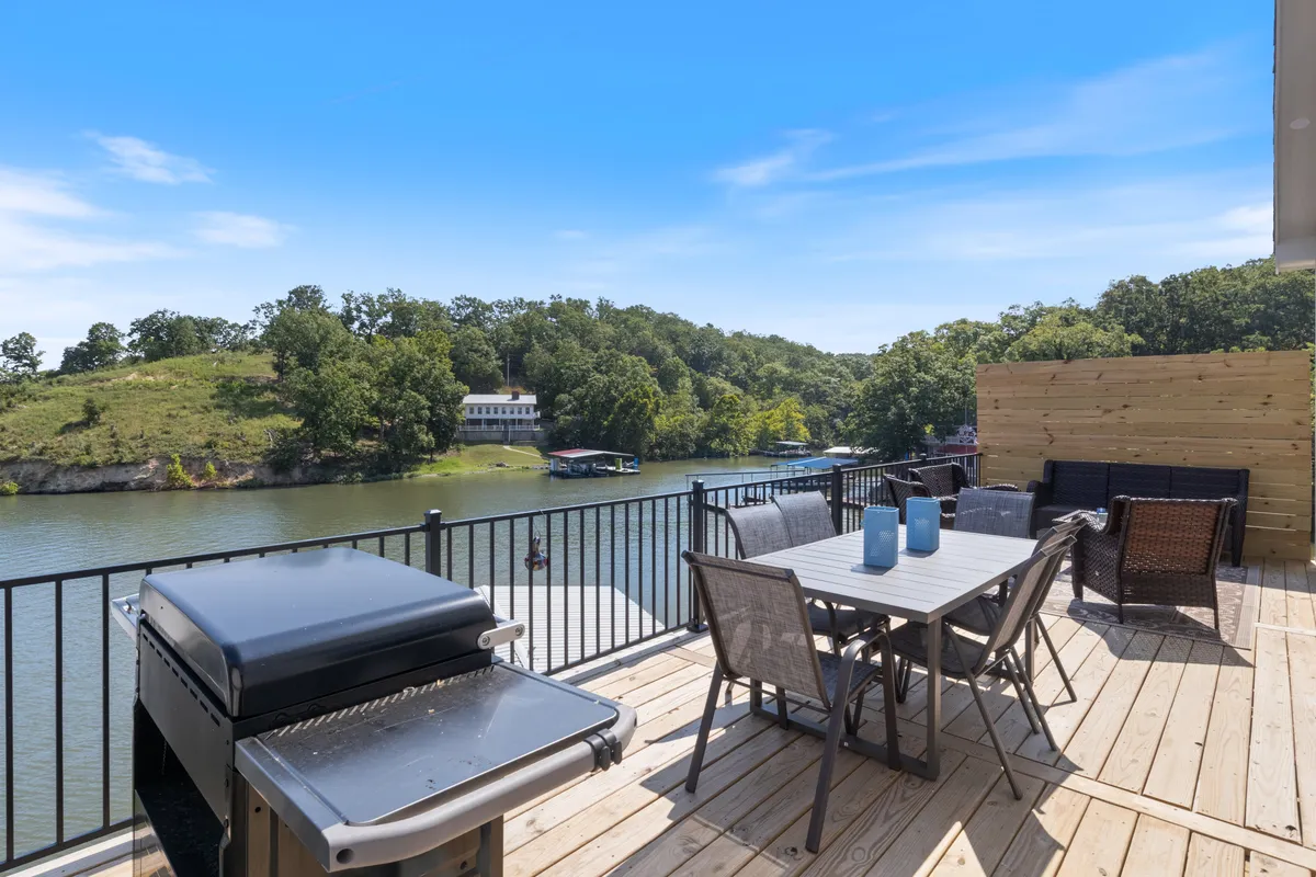 The photo highlights a deck with a grill, dining table, comfortable outdoor seating, and a panoramic view of the lake beyond. The setting suggests an ideal spot for sunset cocktails or morning coffee, with the grill promising memorable barbecues.