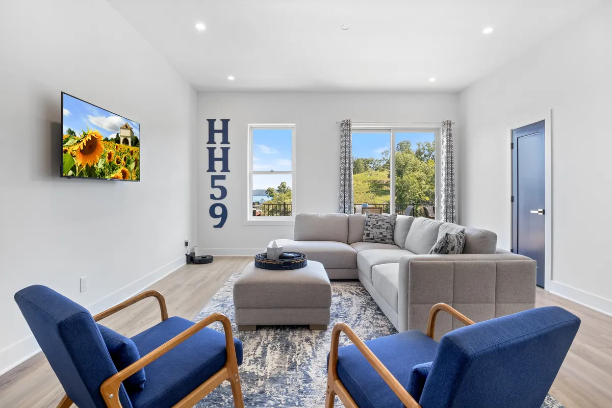 This image features a comfortable living room with a plush sectional sofa, accent chairs, decorative pillows, a wall-mounted television, and large windows that let in copious natural light. The space exudes a welcoming, contemporary vibe and offers a glimpse of the outdoor scenery through the expansive windows.