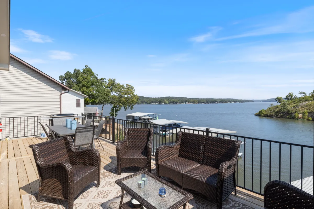 An outdoor seating area and dining table set on a spacious deck overlooking a tranquil lake. The scene features comfortable chairs positioned to maximize enjoyment of the view, with the shimmering water and wooded shoreline visible beyond the railing. The setting suggests both relaxation and opportunities for outdoor dining or social gatherings in a private, scenic environment.