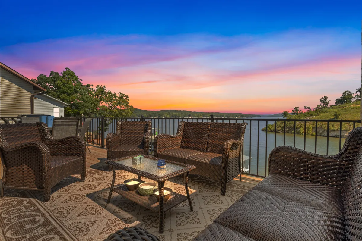 The image features a spacious outdoor seating area overlooking a tranquil lake, with a vibrant sunset painting the sky. Comfortable lounge furniture invites guests to relax, while the private dock and surrounding water highlight direct lake access. The scene exudes serenity, perfect for gathering with friends or family as daylight fades.