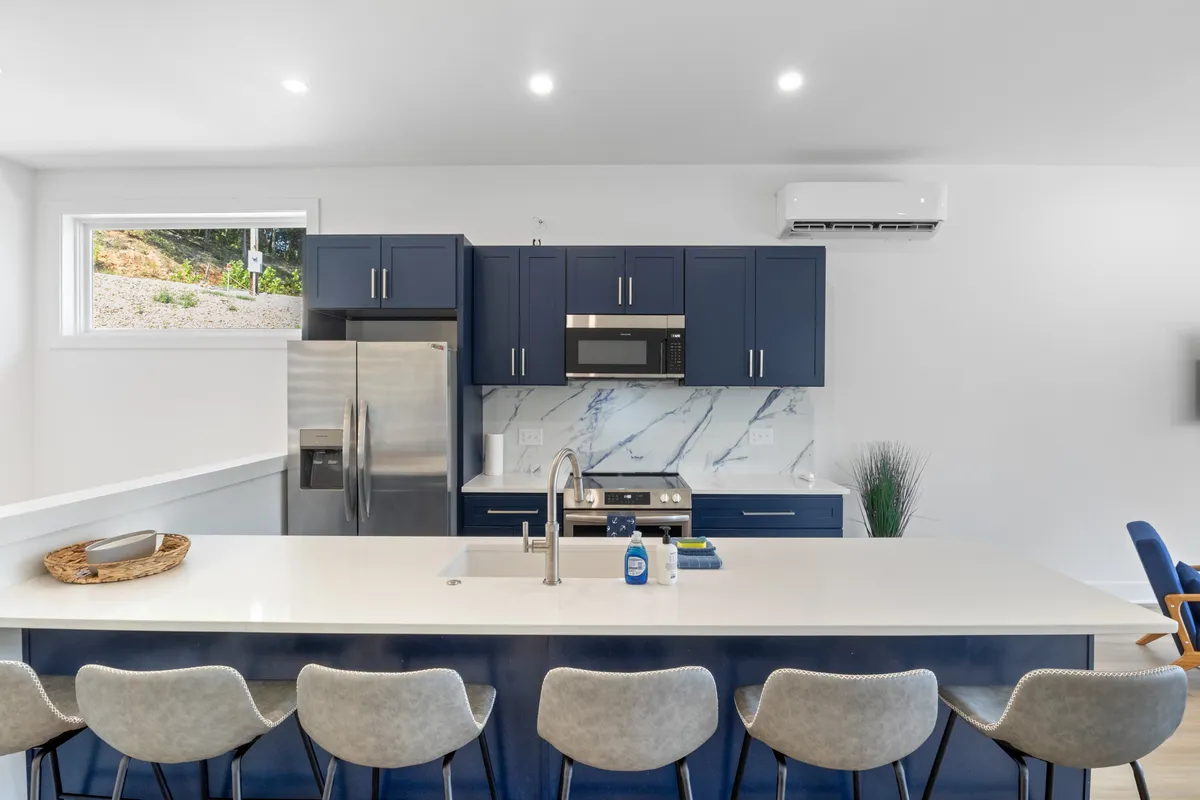 A bright, contemporary kitchen featuring a spacious island with bar seating, gleaming stainless steel appliances, blue cabinetry, and a marble backsplash. The layout encourages gathering and socializing, with plenty of workspace and culinary tools on display.
