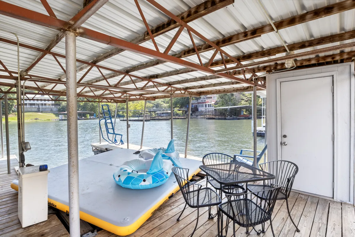 A lively lakeside setup with a floating mat and a blue inflatable dolphin just off the deck. A black outdoor dining set is arranged for meals with a view, and the inviting water backdrop suggests fun swimming and water play for all ages.