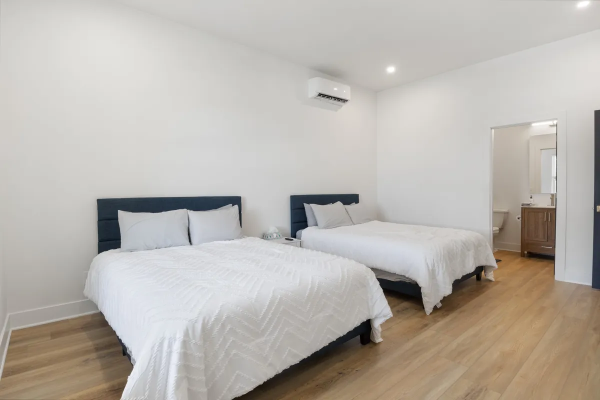 A tranquil bedroom featuring two beds with plush bedding, wooden flooring, and an open doorway that leads to a private bathroom. The soft lighting and serene decor create a restful ambiance, while the layout suggests comfort for couples, families, or friends.