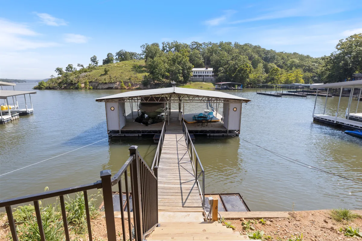 A wooden dock stretches out over calm water, leading to a small boathouse surrounded by lush greenery and distant hills. The dock offers a picturesque spot for boating, fishing, or simply relaxing as the sun sets, with nature enveloping the scene.