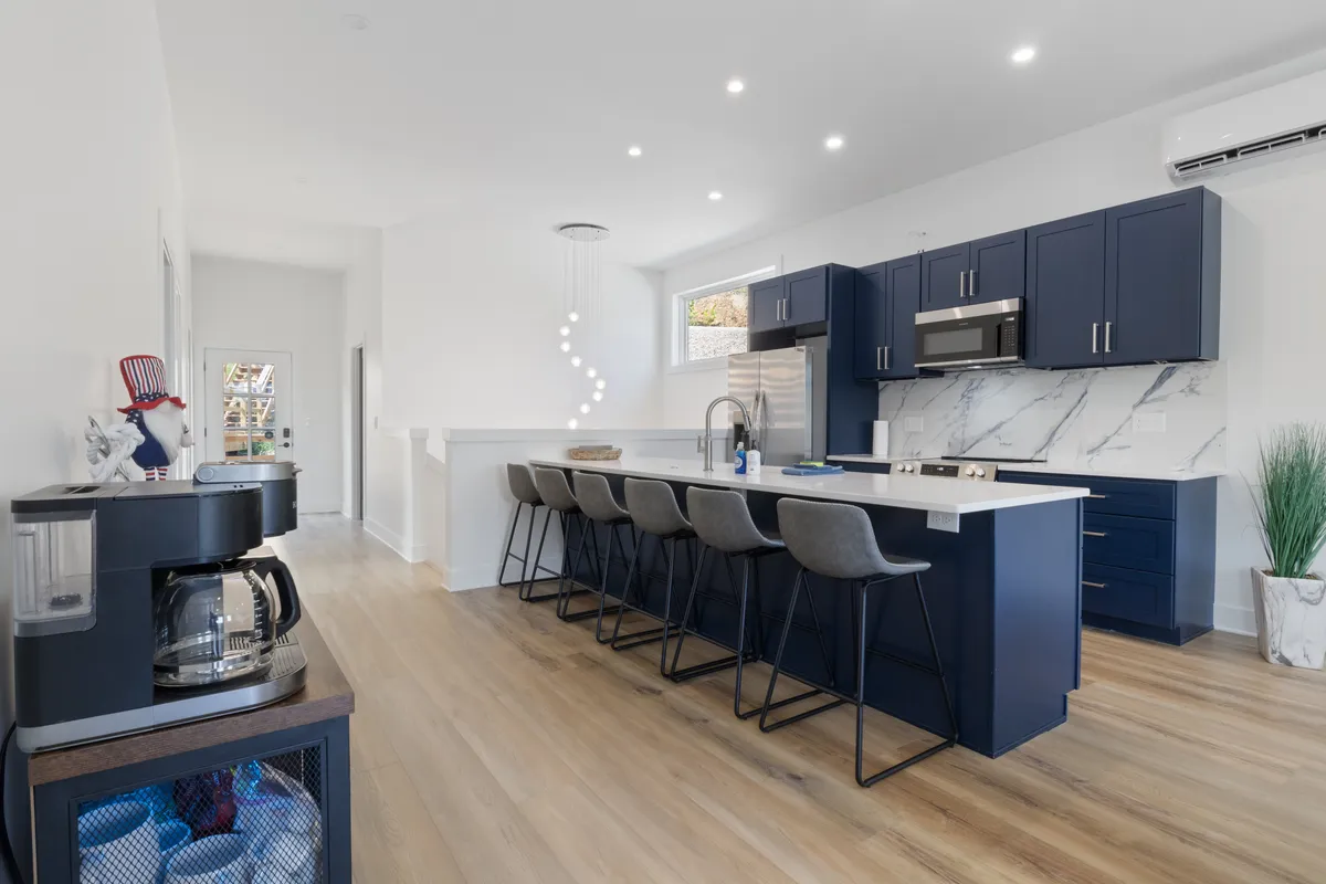 A modern kitchen centered around a large marble-topped island with multiple bar stools, blue cabinetry, and stainless steel appliances. The open design is stylish yet functional, inviting both cooking and conversation.